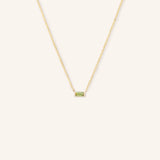 A gold-colored necklace featuring a minimalist baguette design with a peridot gemstone in a white setting.