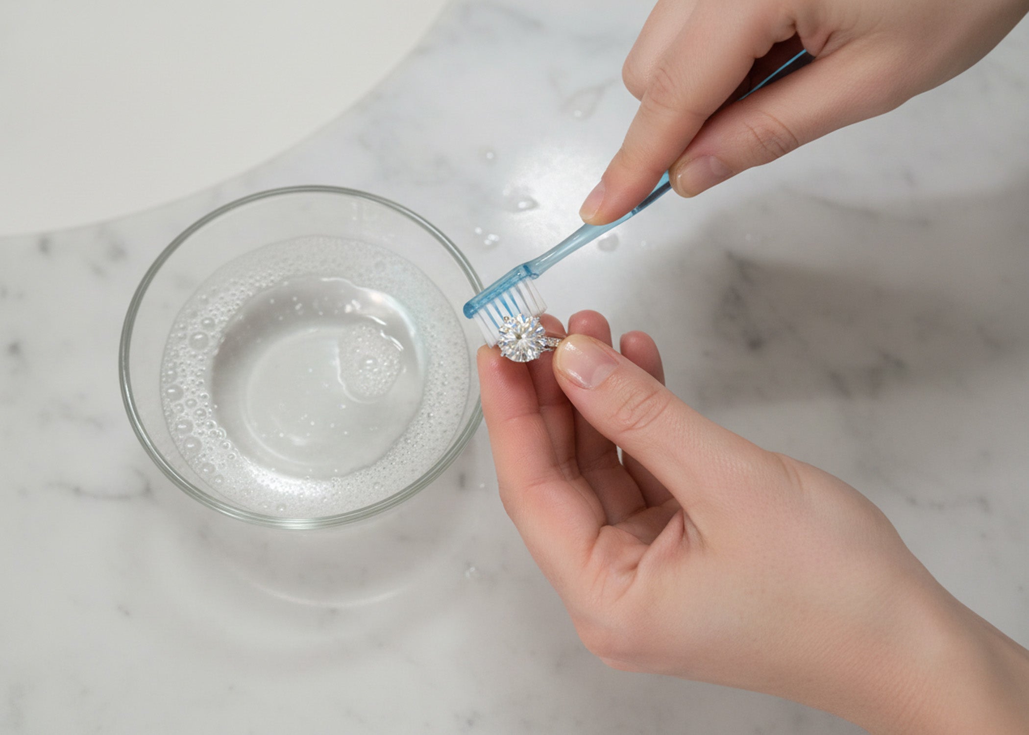 Gold jewelry cleaning setup with bowl of soapy water soft toothbrush and gold pieces on cream linen