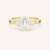 Yellow gold three-stone engagement ring with emerald-cut center diamond and round side diamonds, pave shoulders, on white background