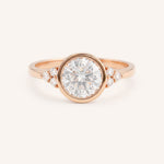 Rose gold round cut bezel set diamond engagement ring with side stones on white background