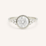 White gold round cut bezel set diamond engagement ring with side stones on white background