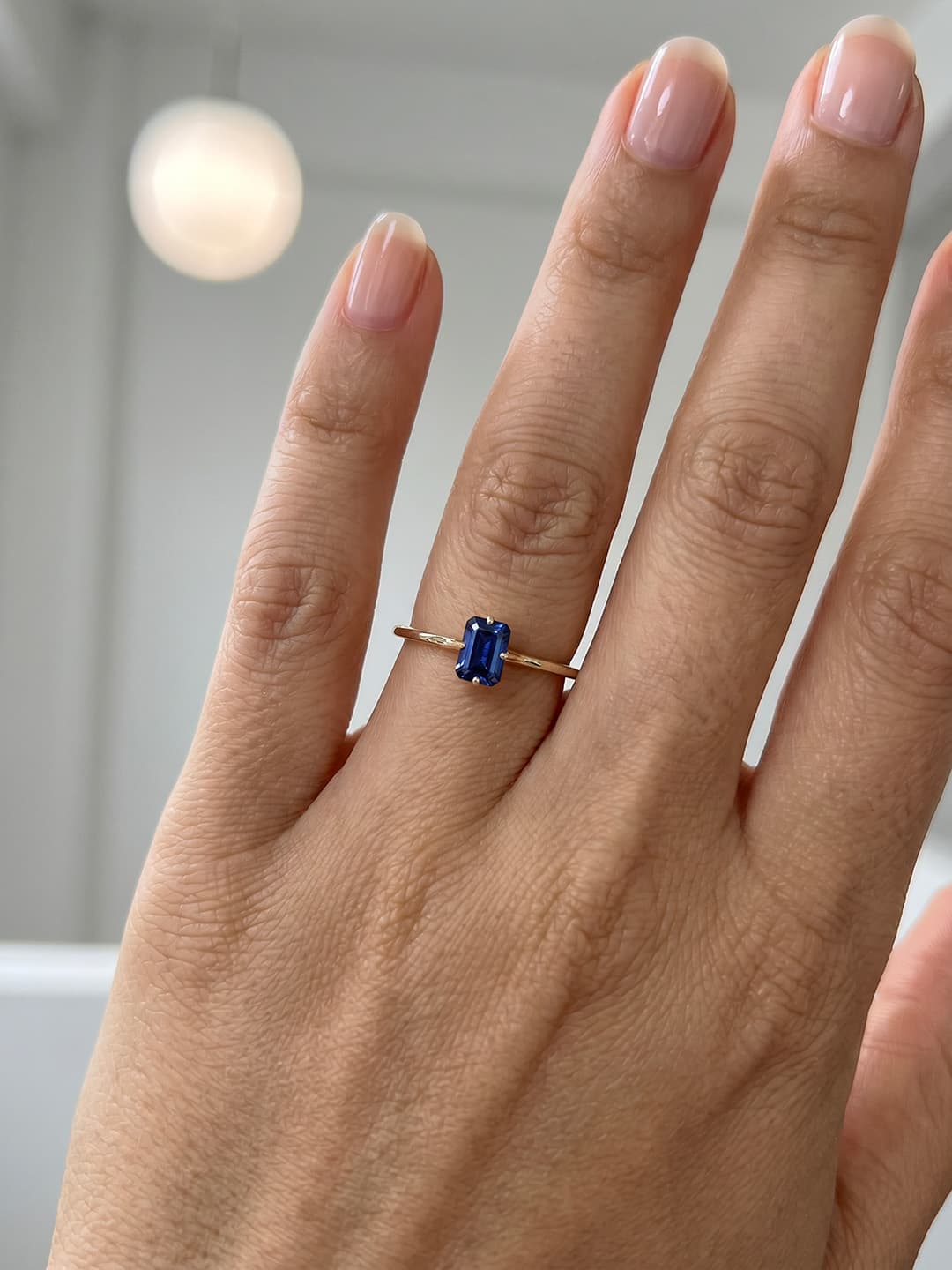 Hand wearing a gold ring with a blue gemstone against a neutral background