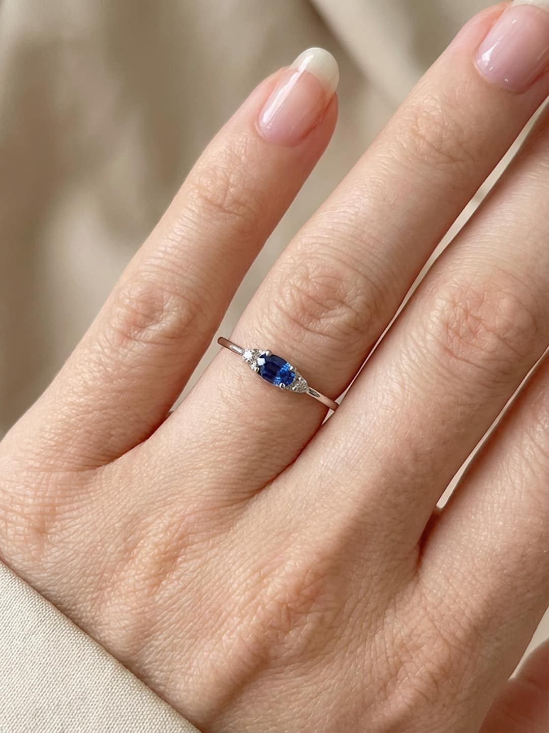 Close-up of a hand wearing a white gold ring with a blue sapphire on a neutral background