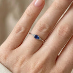 Close-up of a hand wearing a white gold ring with a blue sapphire on a neutral background