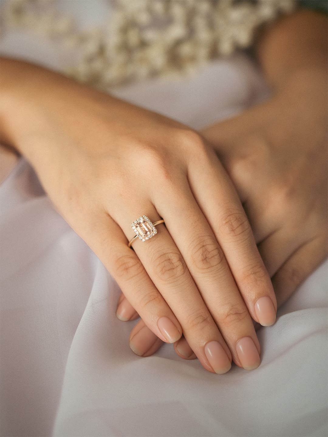 Close-up of a hand wearing a ring with an Emerald Cut morganite and a halo of small diamonds with a soft background