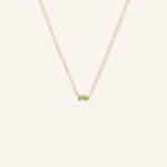 A gold-colored necklace featuring a minimalist baguette design with a peridot gemstone in a white setting.