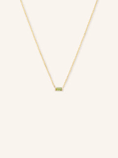 A gold-colored necklace featuring a minimalist baguette design with a peridot gemstone in a white setting.